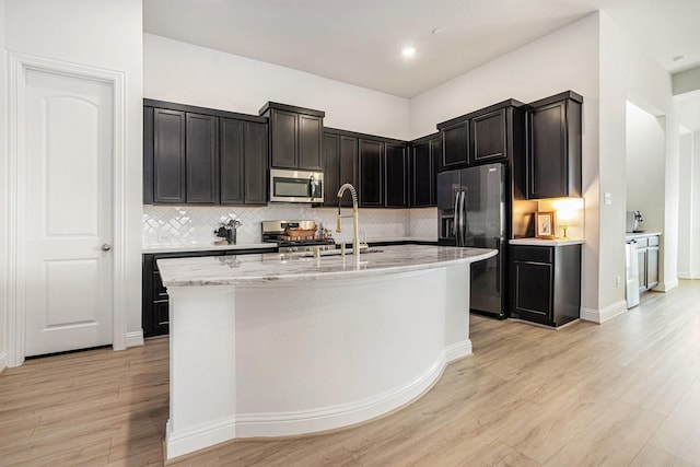 kitchen featuring light stone counters, light wood finished floors, a kitchen island with sink, decorative backsplash, and appliances with stainless steel finishes