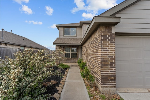 view of exterior entry with brick siding, an attached garage, roof with shingles, and fence