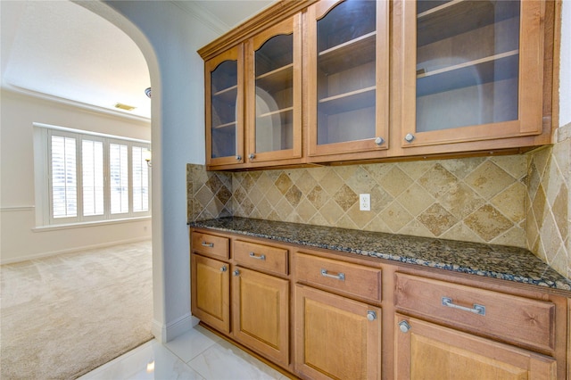 kitchen featuring dark stone counters, arched walkways, glass insert cabinets, light carpet, and backsplash