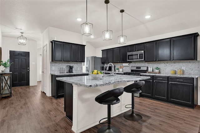 kitchen with light stone counters, dark wood-style floors, a center island with sink, stainless steel appliances, and dark cabinets