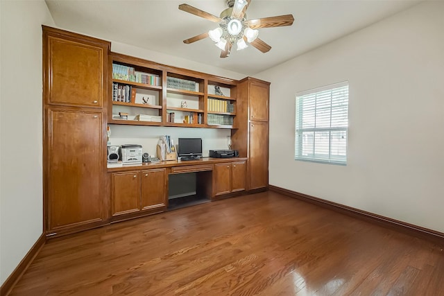unfurnished office with built in desk, baseboards, dark wood-type flooring, and a ceiling fan