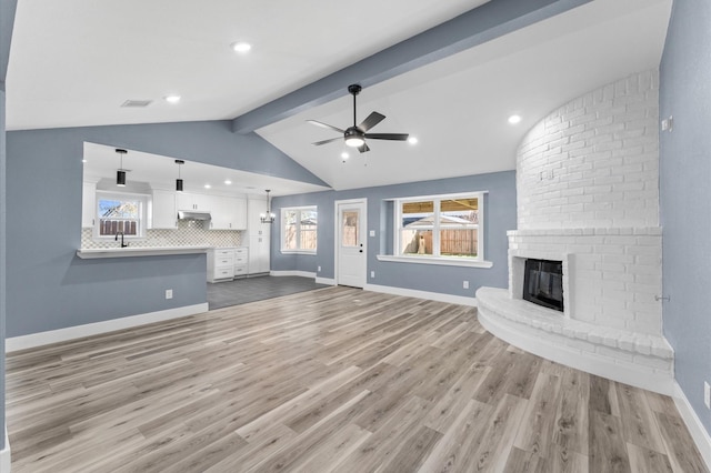 unfurnished living room with a ceiling fan, visible vents, lofted ceiling with beams, light wood-style flooring, and a brick fireplace