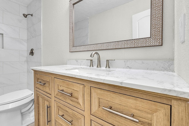 full bath featuring vanity, toilet, a textured wall, and a tile shower