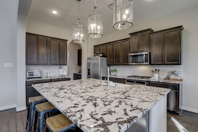 kitchen with a breakfast bar area, an island with sink, arched walkways, stainless steel appliances, and dark brown cabinets