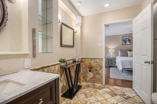 ensuite bathroom featuring connected bathroom, wainscoting, a textured wall, stone tile flooring, and vanity