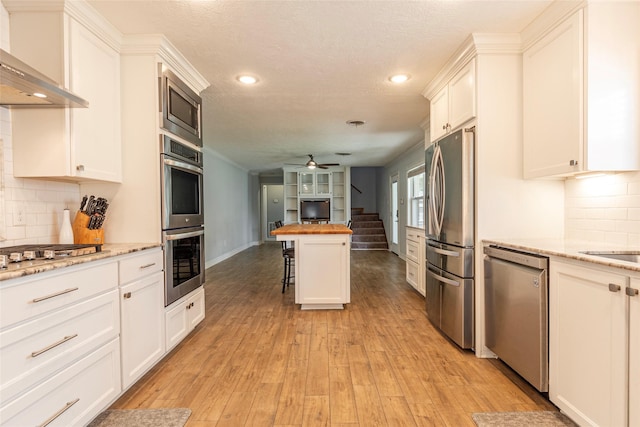 kitchen with wooden counters, ceiling fan, light wood-style floors, appliances with stainless steel finishes, and wall chimney exhaust hood