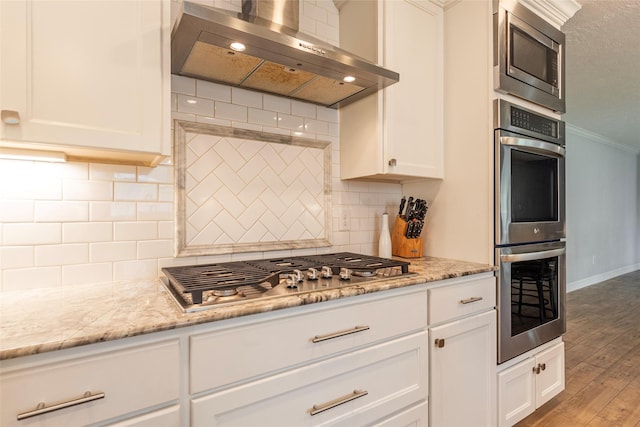 kitchen with wood finished floors, white cabinets, appliances with stainless steel finishes, crown molding, and wall chimney exhaust hood