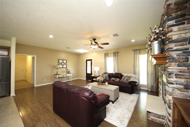 living room featuring a ceiling fan, dark wood finished floors, a stone fireplace, baseboards, and visible vents