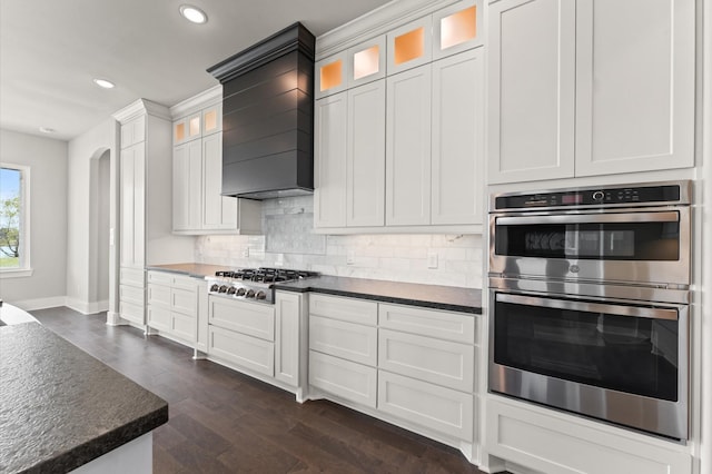 kitchen with wall chimney exhaust hood, stainless steel appliances, tasteful backsplash, dark wood-type flooring, and arched walkways