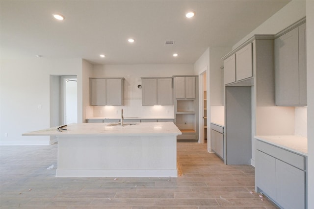 kitchen featuring light wood-style floors, recessed lighting, a kitchen island with sink, a sink, and gray cabinetry