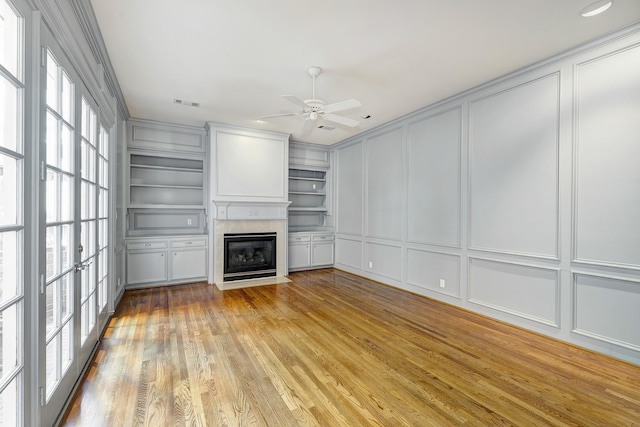 unfurnished living room featuring built in features, a ceiling fan, a fireplace with flush hearth, a decorative wall, and light wood-type flooring