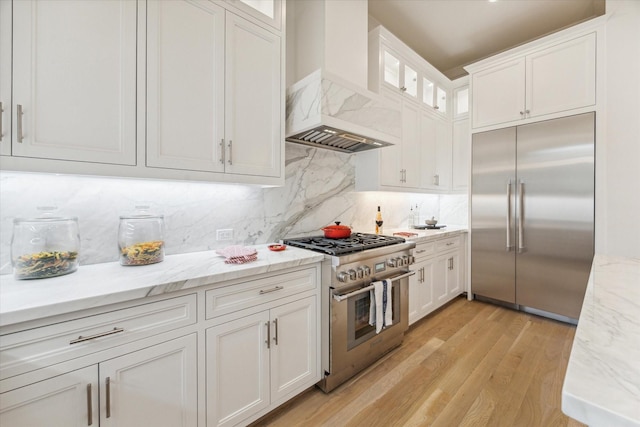 kitchen with light wood-style flooring, tasteful backsplash, premium appliances, white cabinetry, and premium range hood