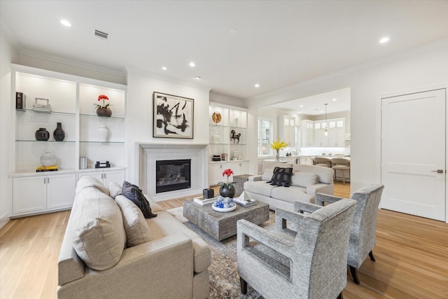 living room with light wood finished floors, a glass covered fireplace, visible vents, recessed lighting, and crown molding