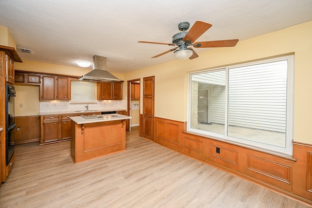 kitchen featuring exhaust hood, light countertops, brown cabinetry, wainscoting, and a kitchen island