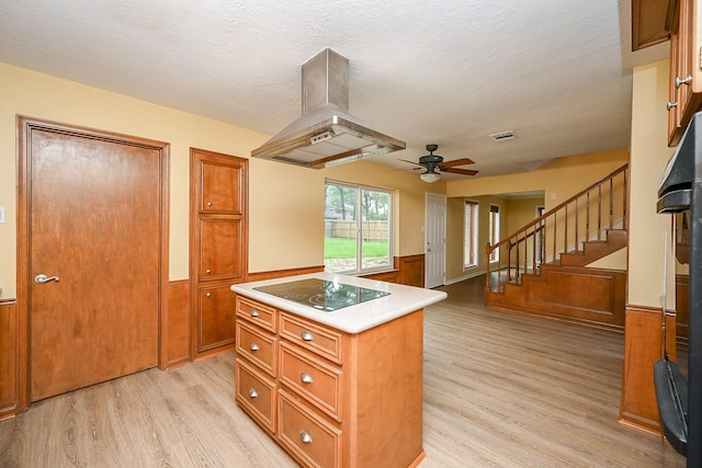 kitchen featuring island exhaust hood, a wainscoted wall, light wood-style floors, brown cabinetry, and black electric cooktop