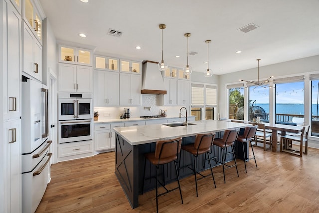 kitchen with visible vents, a sink, white appliances, a kitchen island with sink, and custom range hood