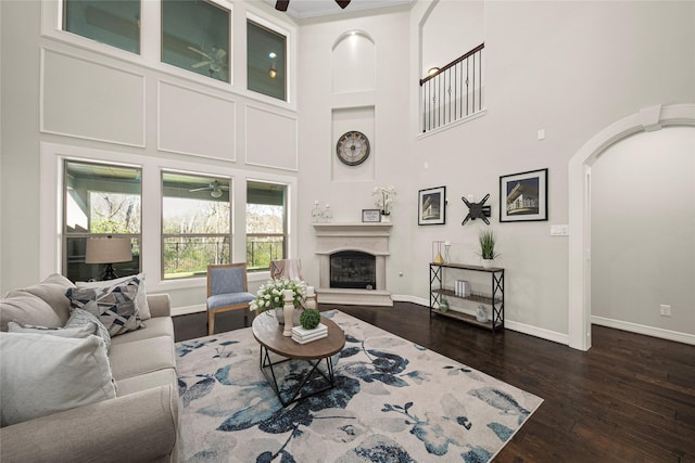 living room featuring baseboards, ceiling fan, a towering ceiling, a glass covered fireplace, and dark wood-style flooring
