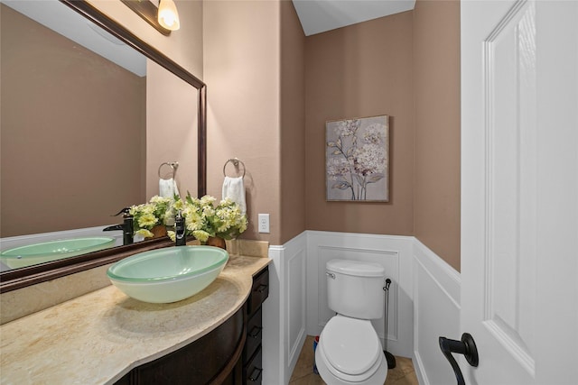 half bathroom with toilet, a decorative wall, wainscoting, tile patterned flooring, and vanity