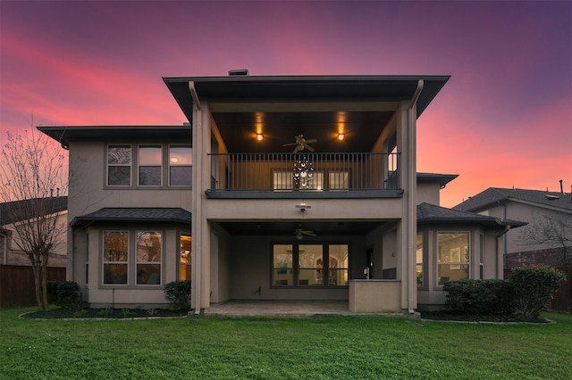 back of property at dusk featuring a balcony, ceiling fan, a lawn, and a patio area