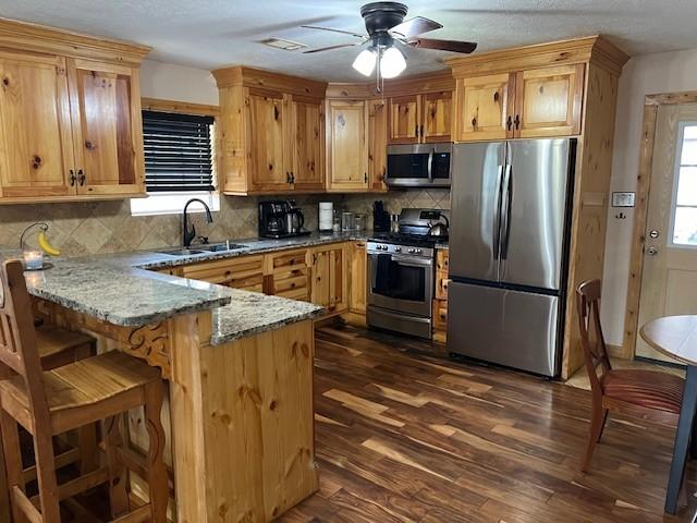kitchen featuring a sink, appliances with stainless steel finishes, a peninsula, stone counters, and dark wood-style flooring