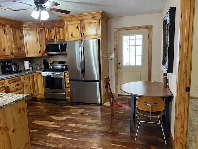 kitchen with backsplash, stone counters, appliances with stainless steel finishes, dark wood-style floors, and a ceiling fan