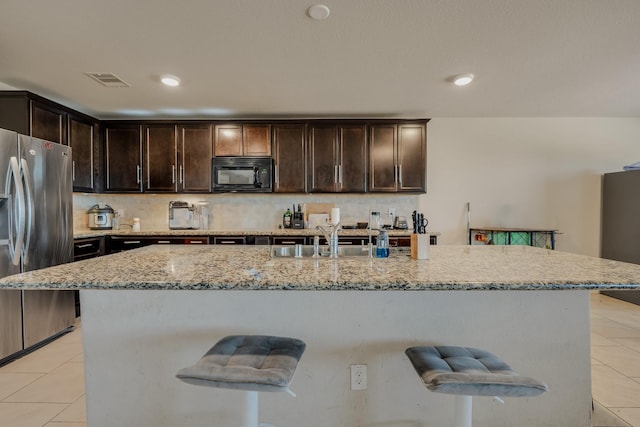 kitchen with black microwave, dark brown cabinetry, a breakfast bar area, light tile patterned flooring, and stainless steel fridge