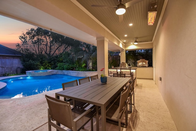 pool at dusk featuring outdoor dining space, a patio, a ceiling fan, fence, and an outdoor kitchen