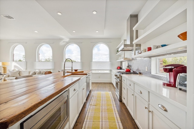 kitchen with butcher block counters, wall chimney range hood, open shelves, a sink, and stainless steel appliances