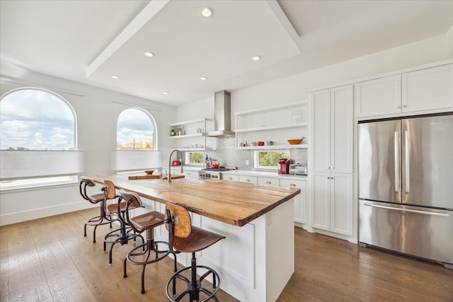 kitchen with open shelves, a sink, wall chimney range hood, a center island with sink, and stainless steel appliances
