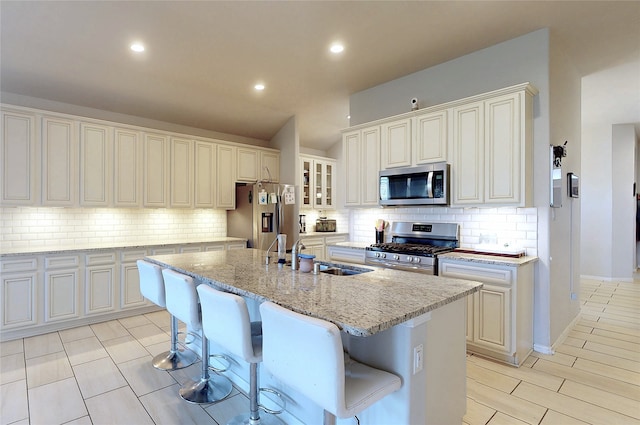 kitchen featuring a kitchen island with sink, tasteful backsplash, a sink, light stone counters, and stainless steel appliances