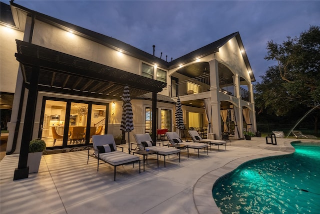 back of house at night with an outdoor pool, stucco siding, a balcony, and a patio area