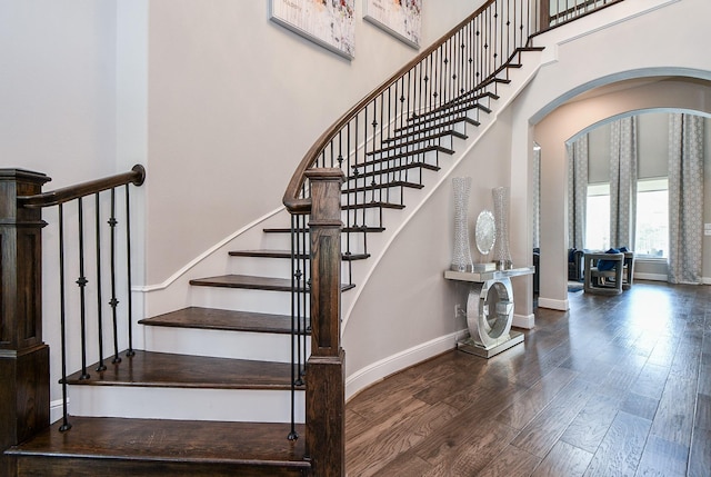 staircase featuring arched walkways, a high ceiling, baseboards, and wood finished floors