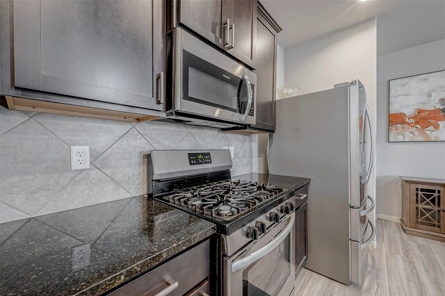 kitchen featuring tasteful backsplash, light wood finished floors, dark brown cabinets, dark stone counters, and appliances with stainless steel finishes