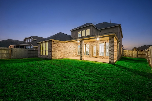 back of house at dusk with a lawn, a fenced backyard, brick siding, and a patio area