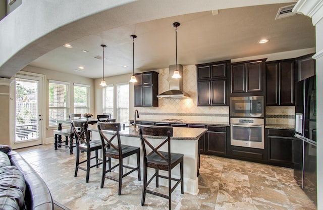 kitchen with a breakfast bar, backsplash, arched walkways, appliances with stainless steel finishes, and wall chimney exhaust hood