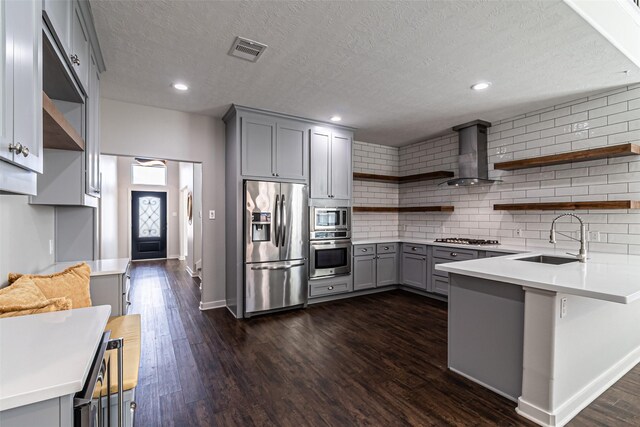 kitchen with open shelves, gray cabinets, a sink, appliances with stainless steel finishes, and wall chimney range hood