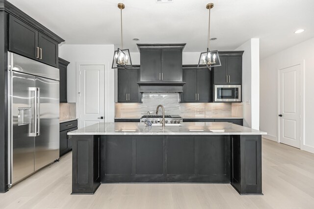 kitchen with tasteful backsplash, an inviting chandelier, light wood-style floors, an island with sink, and built in appliances