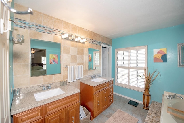 ensuite bathroom with tile patterned floors, two vanities, a ceiling fan, a bath, and tile walls
