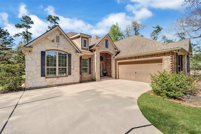 french country style house featuring stone siding, an attached garage, brick siding, driveway, and a shingled roof