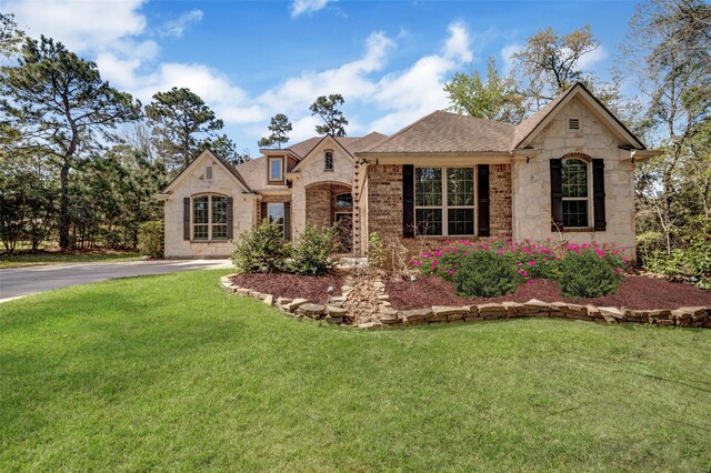 french country inspired facade featuring stone siding, a front yard, brick siding, roof with shingles, and driveway