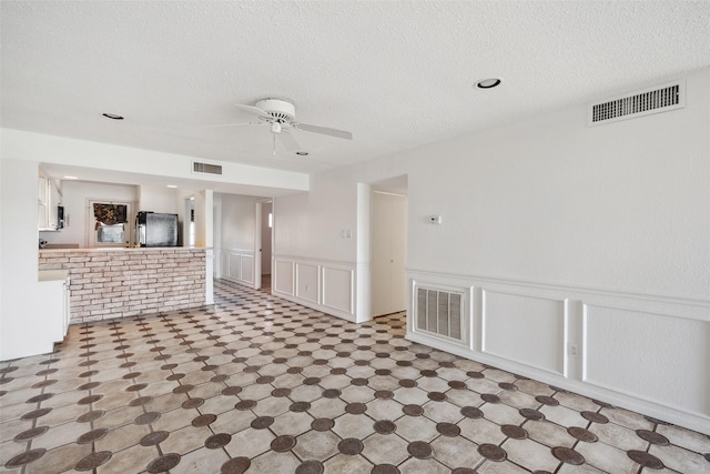 unfurnished living room featuring ceiling fan, a decorative wall, wainscoting, and a textured ceiling