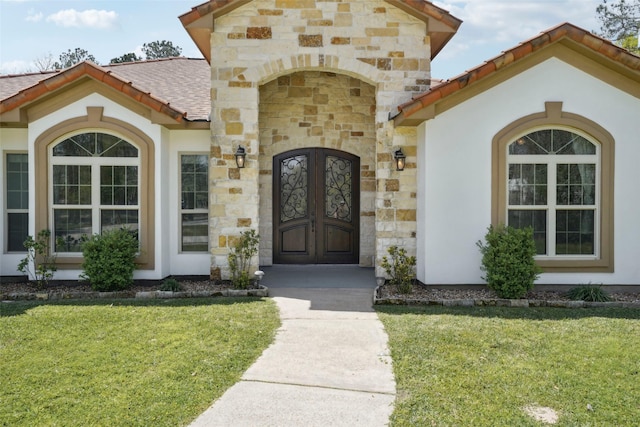 view of exterior entry featuring roof with shingles, a yard, stucco siding, and stone siding
