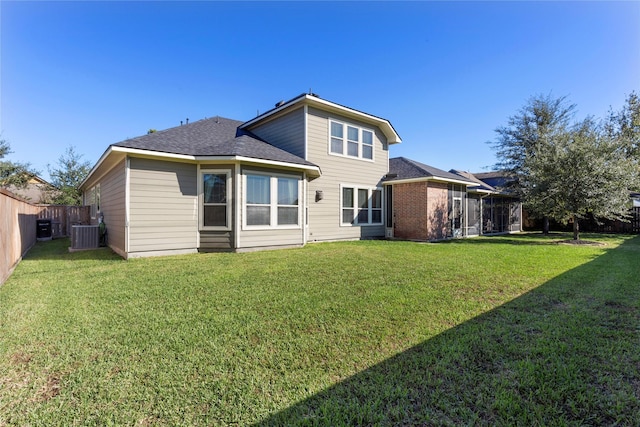 rear view of house featuring a lawn, fence, central AC unit, and roof with shingles