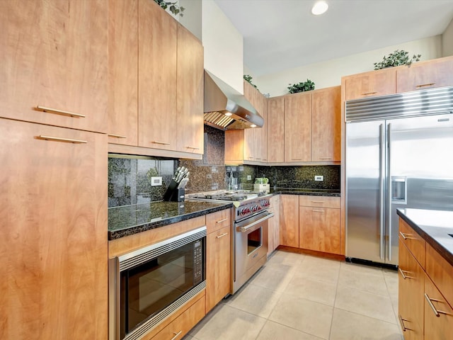 kitchen featuring light tile patterned floors, backsplash, dark stone counters, built in appliances, and wall chimney exhaust hood