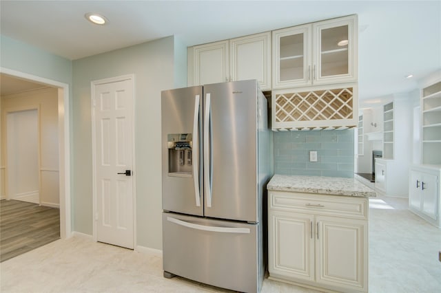 kitchen featuring baseboards, glass insert cabinets, cream cabinetry, light stone counters, and stainless steel fridge with ice dispenser