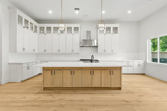 6536 Community Dr, West University Place, TX 77005 - kitchen featuring wall chimney exhaust hood, light countertops, a center island with sink, light wood-style floors, and glass insert cabinets