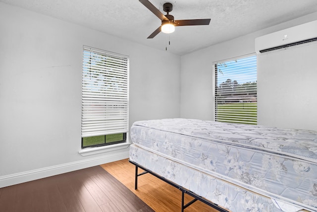 bedroom featuring wood finished floors, baseboards, a wall unit AC, a ceiling fan, and a textured ceiling