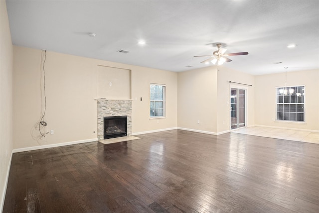 unfurnished living room with hardwood / wood-style floors, ceiling fan with notable chandelier, baseboards, visible vents, and a stone fireplace