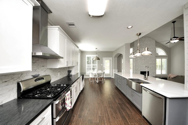 kitchen with a sink, wall chimney exhaust hood, stainless steel appliances, ceiling fan with notable chandelier, and visible vents