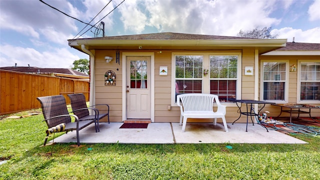view of exterior entry with fence, roof with shingles, a patio, and a lawn